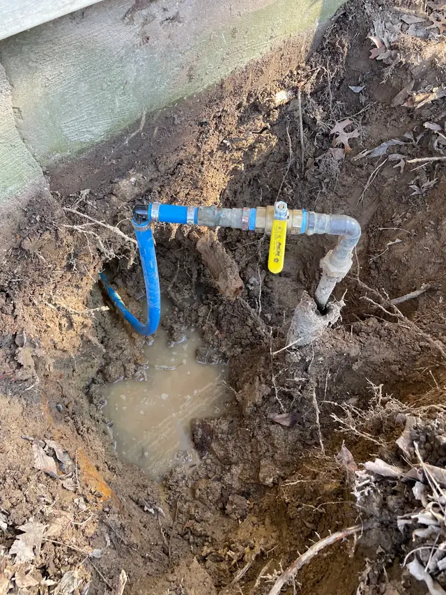 A main water line dug out beside a foundation, blue PEX and a yellow ball valve over the leak